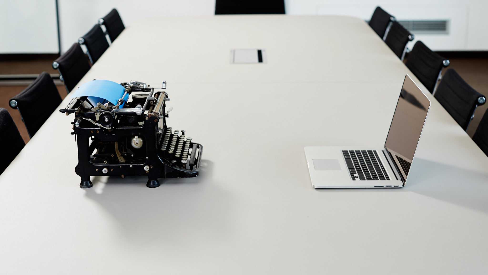 A modern laptop sits opposite an old typewriter on a conference table.